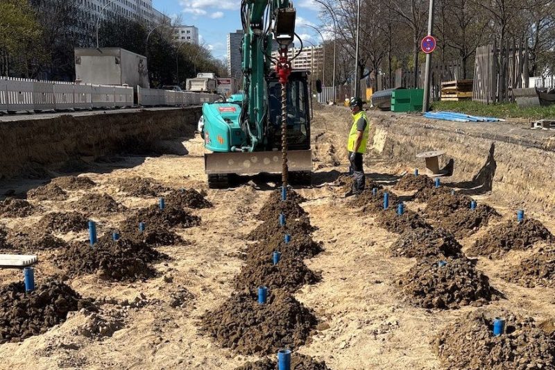 bohrlochsondierung-kampfmittel-sondierung-baustelle-berlin Durchführung einer Bohrlochsondierung zur Kampfmittelüberprüfung auf einer innerstädtischen Baustelle in Berlin. Ein Bagger mit Bohrgerät erstellt Sondierbohrungen, markiert durch blaue Kunststoffrohre, zur Untersuchung des Untergrunds auf mögliche Kampfmittelfunde.